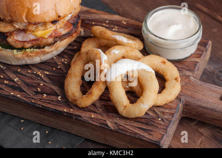 Nahaufnahme von gebratenen Zwiebelringen auf dem Brett, mit einer weißen Knoblauchsauce auf Holzhintergrund. Stockfoto