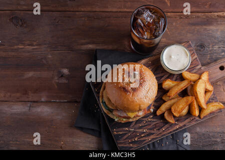 Ein leckerer Burger mit Snacks in Form von Kartoffeln mit weißer Knoblauchsauce und einem Glas Cola. Saftiger Burger mit Zwiebelringen, Speck und Käse o Stockfoto