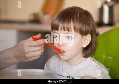 Schönes Baby isst Brei von Mama's Hand. Die Mutter füttert, der Sohn mit einem Löffel. Junge mit langen blonden Haaren und großen blauen Augen wahrscheinlich. Stockfoto