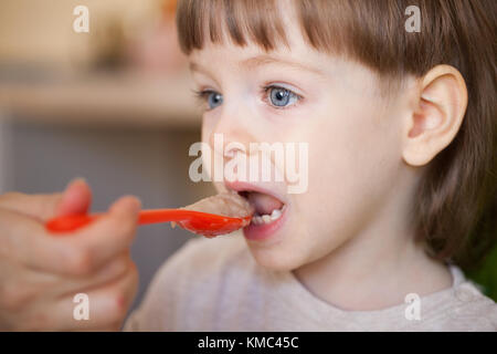 Schönes Baby isst Brei von Mama's Hand. Die Mutter füttert, der Sohn mit einem Löffel. Junge mit langen blonden Haaren und großen blauen Augen wahrscheinlich. Stockfoto