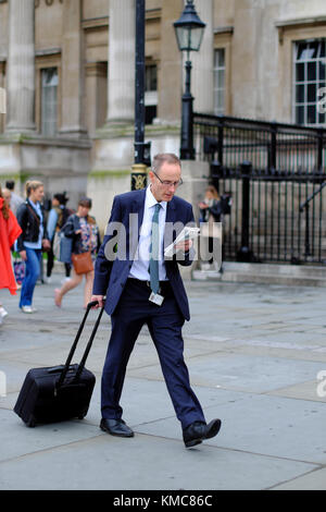 Kaukasische Mann ziehen Reisetasche und lesen Zeitung, während man durch den Trafalgar Square, London, England, Großbritannien Stockfoto
