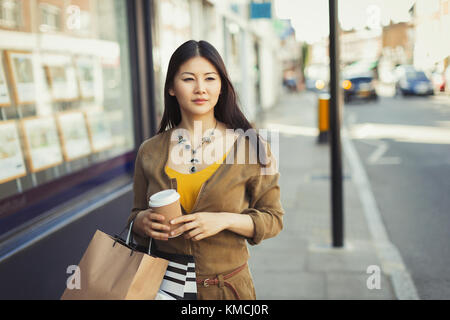 Junge Frau, die mit einer Kaffeetasse und einem Einkaufsbummel durch das Schaufenster läuft Taschen Stockfoto