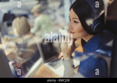 Nachdenkliche junge Frau am Laptop, die mit Kopfhörern Musik hört Am Cafe-Fenster Stockfoto