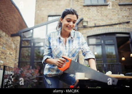 Junge Frau mit Säge, die Holz auf der Terrasse geschnitten hat Stockfoto