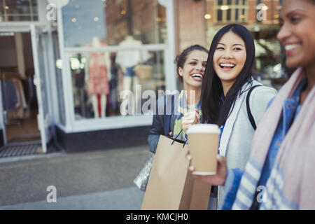 Lachende Freundinnen, die mit Kaffee und Einkaufen entlang der Ladenfront gehen Taschen Stockfoto