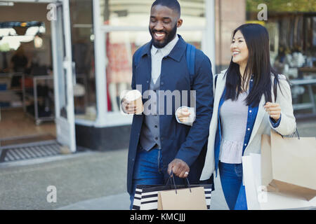 Lächelndes junges Paar, das Arm in Arm entlang der Schaufenster mit geht Kaffee und Einkaufstaschen Stockfoto