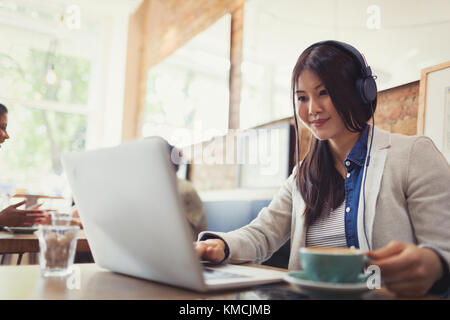 Lächelnde junge Frau, die mit Kopfhörern am Laptop Musik hört Und Kaffee trinken im Café Stockfoto