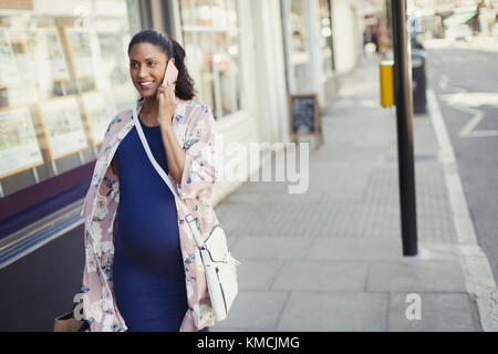 Lächelnde schwangere Frau, die mit dem Handy spricht und durch die Stadt läuft Stockfoto