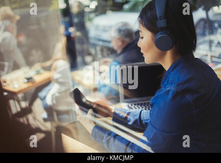 Junge Frau hört Musik mit Kopfhörern, SMS mit Handy am Cafe-Fenster Stockfoto