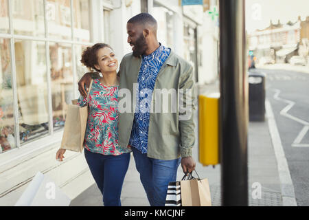 Liebevolles junges Paar mit Einkaufstaschen, die entlang der städtischen Schaufenster laufen Stockfoto