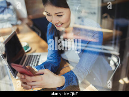 Lächelnde junge Frau, die mit Kopfhörern und SMS Musik hört Mit Handy am Laptop im Cafe Fenster Stockfoto