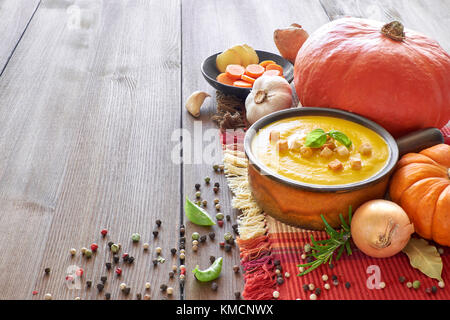 Kürbis, Karotten und Ingwer creme Suppe in Keramik Topf auf dem Holztisch, gewürzt mit Croutons und Basilicum. Zutaten: Kürbis, Karotten, Ingwer Stockfoto