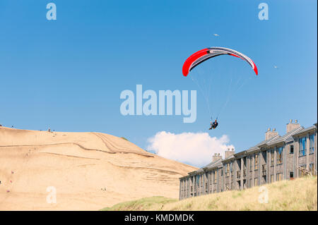 Ein Hängegleiter fliegt über ein Gebäude bei Pacific City Oregon. Sand Hügel im Hintergrund mit blauem Himmel und weißen Wolken. Stockfoto