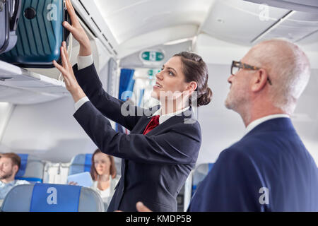 Der Flugbegleiter hilft dem Geschäftsmann, das Gepäck in den Gepäckraum zu legen Flugzeug Stockfoto