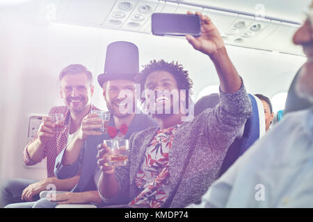 Enthusiastische junge männliche Freunde mit Kamera-Handy trinken und nehmen Selfie im Flugzeug