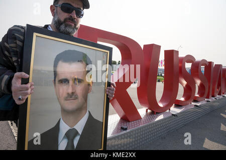 Der Mann hält ein Porträt von Präsident Baschar Assad in der Nähe der Inschrift "Russland 2018" im Olympischen Park von Sotschi, die Region Krasnodar, Russland Stockfoto