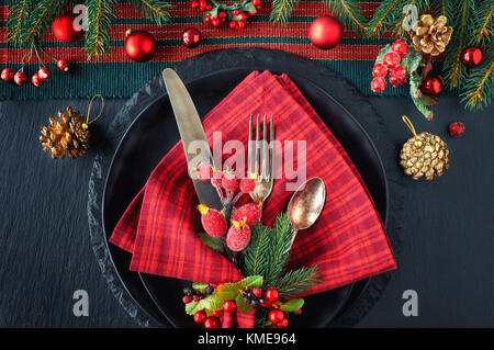 Weihnachtsmenü Konzept auf dunklem Hintergrund. Die schwarzen Platten und vintage Besteck mit Weihnachtsschmuck in Grün und Rot. Stockfoto