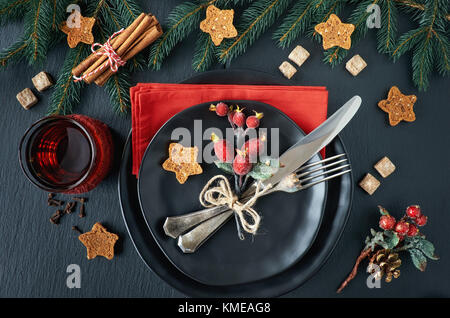 Weihnachtsmenü Konzept auf dunklem Hintergrund. Die schwarzen Platten und vintage Besteck mit Weihnachtsschmuck in Grün und Rot. Stockfoto