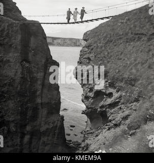 1950, historische, ein Mann und zwei Damen für ein Bild, das auf der berühmten pose Carrick-a-Rede rope bridge, Co Antrim, Nordirland. Die hängebrücke ist fast 100 m hohen verbindet das Festland zu der kleinen Insel Carrickarede und wurde erstmals im Jahr 1755 von lokalen Lachs Fischer gegründet. Stockfoto