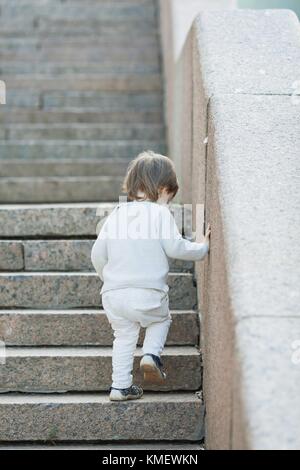 Kleiner blauäugiger Junge mit langen Haaren klettert die Steintreppe hinauf. Das Konzept des Aufwachsens. Stockfoto