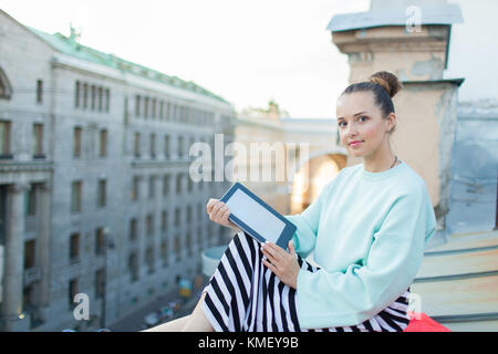 Attraktive und stilvolle Mädchen sitzt auf dem Dach des Hauses in der Altstadt und liest das E-Book. Stockfoto