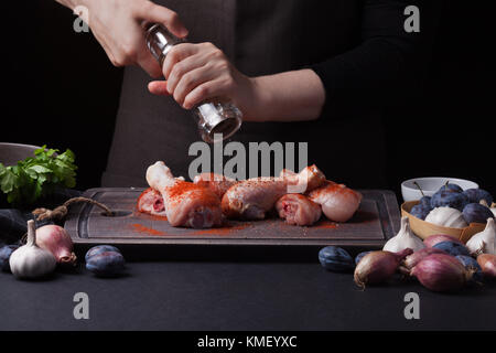 Eine Köchin streut frische rohe Hähnchendrumknüppel auf dunklem Hintergrund mit einer Mischung aus Paprika. In der Nähe liegen die Zutaten zum Kochen: Schalotten, Stockfoto