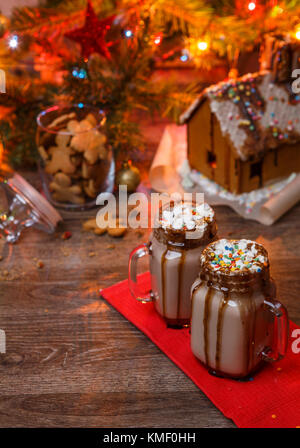 Zwei Gläser Kakao mit Marshmallows, Sahne und Schokolade Sirup auf Holztisch und Lebkuchenhaus, Cookie Jar und Weihnachtsbaum mit Spielzeug und Girlande für den Hintergrund. Stockfoto