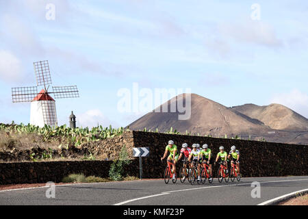 Gruppe von Radfahrern Radeln auf der Straße mit Vulkanen und Windmühle im Hintergrund, Lanzarote, Kanarische Inseln, Spanien Stockfoto