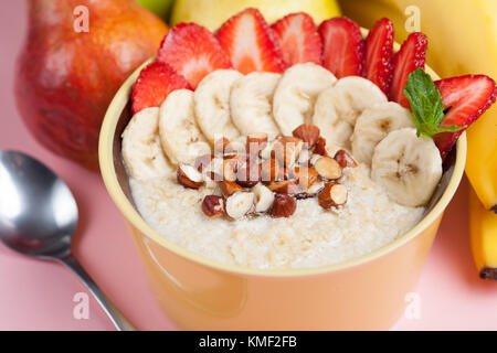 Schüssel Haferflocken mit einer Banane, Erdbeeren, Mandeln, Haselnüssen und Butter auf einem rosa Hintergrund. Warmes und gesundes Gericht zum Frühstück, Draufsicht. Stockfoto