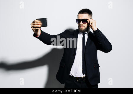 Verspielte Macho in Schwarz classic Anzug und Sonnenbrille unter selfie auf dem Smartphone, auf weißem Hintergrund Stockfoto