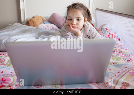 Kleines Mädchen im Bett liegen und Filme mit einem Laptop in seinem Schlafzimmer. Sie schaut gelangweilt Stockfoto