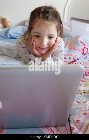 Kleines Mädchen im Bett liegen und spielen mit einem Laptop in seinem Schlafzimmer. Sie sieht Spaß Stockfoto