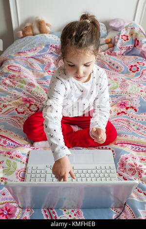 Kleines Mädchen im Bett sitzen und spielen online Spiele mit Laptop. Sie ist durch Berühren des Bildschirms Stockfoto