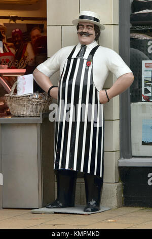 Eine große lebensgroße Modell oder Büste eines Metzgers tragen ein Stroh Kreissäge Hut und ein blau-weißes traditionelles gestreiften Schürze außerhalb von einem Metzger Shop. Stockfoto