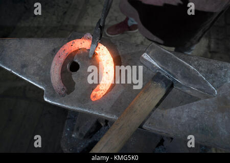 Lehrling des Schmieds, der in der Werkstatt ein rotes, heißes Hufeisen auf einen Amboss schlägt Stockfoto