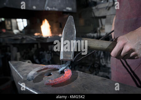 Lehrling des Schmieds, der in der Werkstatt ein rotes, heißes Hufeisen auf einen Amboss schlägt Stockfoto