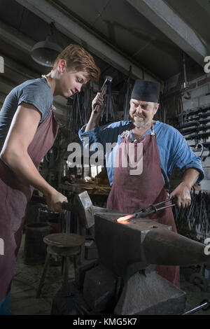 Schmied und Lehrling hammerten in der Werkstatt eine rote Bar mit heißem Eisen Stockfoto