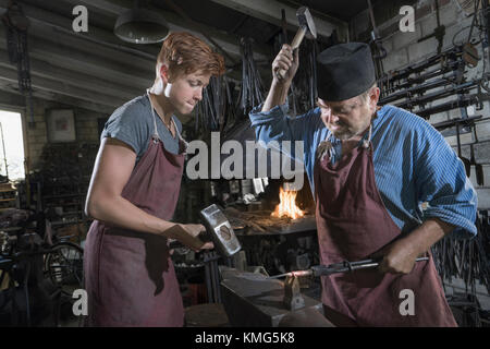 Schmied und Lehrling hammerten in der Werkstatt eine rote Bar mit heißem Eisen Stockfoto