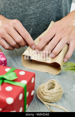 Nahaufnahme eines jungen kaukasischen Mann ein Geschenk auf einem grauen rustikalen Tisch voller Geschenke in Papiere mit unterschiedlichen Mustern gewickelt und mit Bändern gebunden Stockfoto