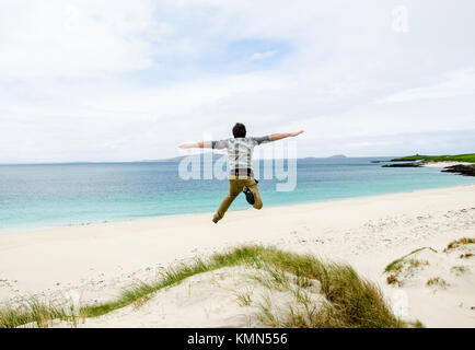 Junge Modell springen auf einer Düne mit offenen Armen. Weißer Sandstrand und blauen Himmel im Hintergrund. Schottland, Großbritannien. Stockfoto