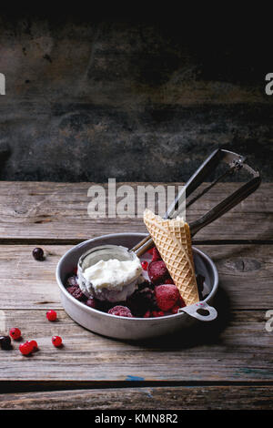 Aluminium-Platte mit Vanilleeis, Mischung aus gefrorenen Beeren und gebissen Waffer Kegel, serviert mit Metalllöffel über Holztisch Stockfoto