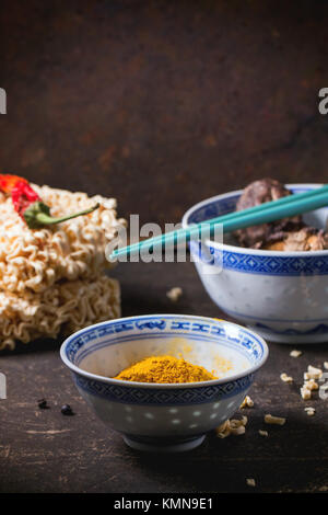 Trockene Nudeln für Ramen Suppe mit kurkuma Pulver und Shiitake-pilze in Porzellan Schalen, mit red hot chili peppers über dunklen Tisch serviert. Siehe Serie Stockfoto