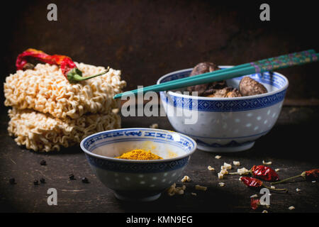 Trockene Nudeln für Ramen Suppe mit kurkuma Pulver und Shiitake-pilze in Porzellan Schalen, mit red hot chili peppers über dunklen Tisch serviert. Siehe Serie Stockfoto