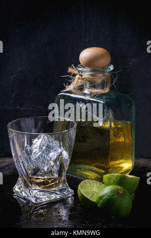 Glas Tequila Anejo mit Eiswürfeln und eine Flasche Tequila, serviert mit in Scheiben geschnittenen Limetten auf schwarzem Hintergrund Stockfoto