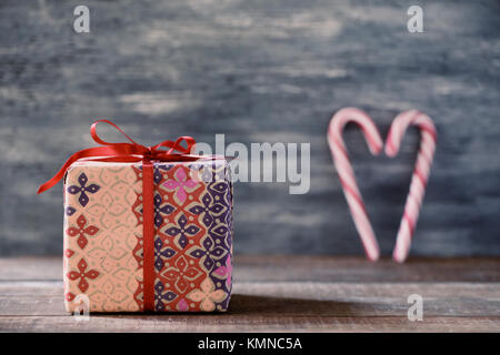 Eine gemütliche Geschenk in einer schönen Papier gewickelt und gebunden mit einem roten Band, und zwei typischen rot-weißen Zuckerstangen bilden ein Herz, auf einem rustikalen Holzmöbeln Oberfläche Stockfoto