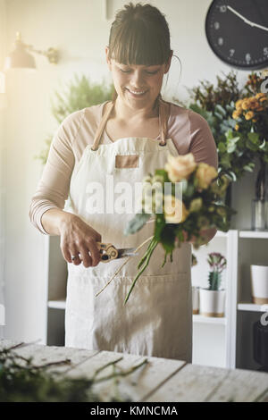 Lächelnde junge weibliche Blumenhändler, der an einem Tisch in Ihrem Blumenladen trimmen stammt und einen Blumenstrauß aus gemischten Blumen Stockfoto