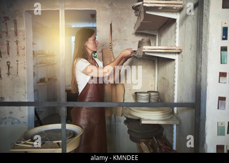 Weibliche Handwerker stehen allein in Ihrer kreativen Keramik Studio, ein frisch gedreht Topf aus Ton auf ein Regal gemacht Stockfoto