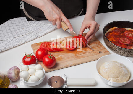 Frau Koch schneidet die Tomaten für die Herstellung hausgemachte Pizza. Auf dem weißen Tisch sind Mozzarella, Pizza Teig, und Knoblauch. Stockfoto