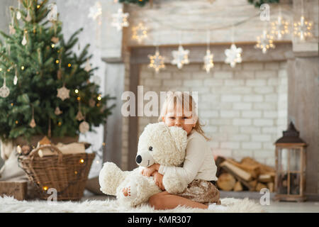 Kind, Mädchen sitzt auf dem Boden, Lächeln und Umarmungen Weißer Teddybär gegen den Hintergrund der Weihnachtsbaum und Dekorationen. Stockfoto