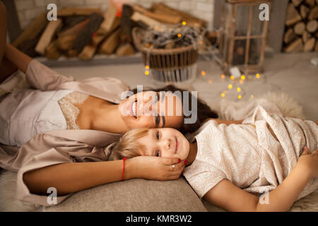 Glückliche Mutter in peignoir mit ihrer kleinen Tochter liegt auf dem Boden und lächelt. Von der Seite. Stockfoto